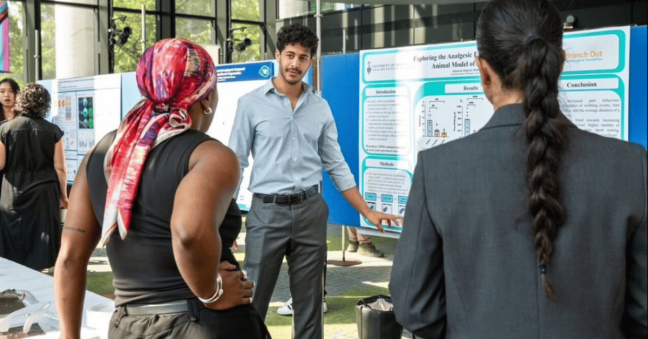 An image of a young male scientist pointing to a board as he presents a research poster at a science conference